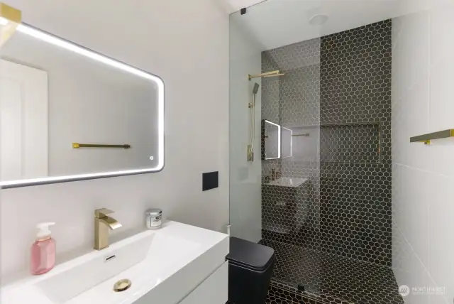 Main level bathroom with floor to ceiling tiled walls and shower with dual heads. Lighted mirror and wall mounted toilet.