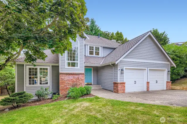 Well-maintained two-story home with gray siding, brick accents, and a double-car garage. Mature trees and landscaped yard offer natural shade and privacy.