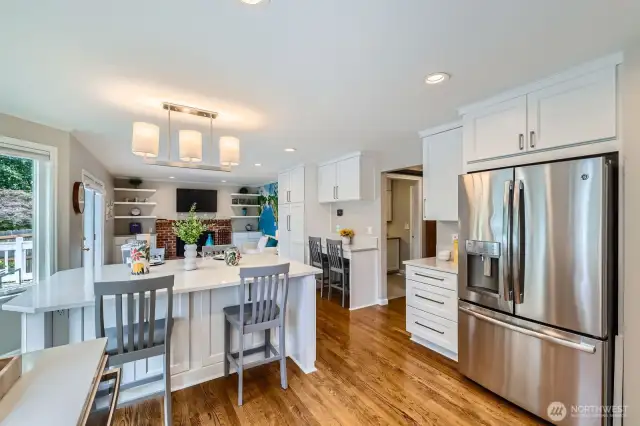 Open kitchen layout with large island, breakfast bar seating, and built-in workspace. Overlooks the family room with brick fireplace and custom shelving.