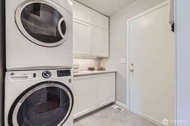 Crisp and efficient laundry space with stacked washer/dryer, upper and lower cabinetry, and tile flooring. Convenient access to the garage.
