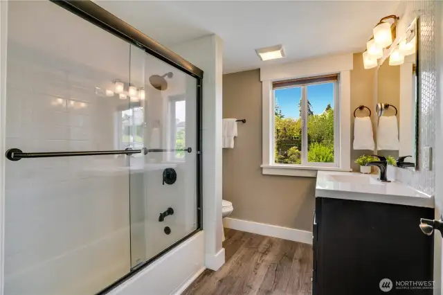 The primary bathtub and shower feature a full subway tile surround and classic glass doors. The vanity and backsplash harmonize with the other bathrooms in the home, while the luxury vinyl flooring ensures easy maintenance.