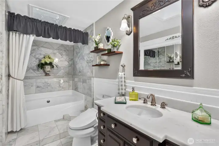 “Marble bathroom featuring a large soaking tub, wainscoting, and two skylights.” “Marble bathroom featuring a large soaking tub, wainscoting, and two skylights.”