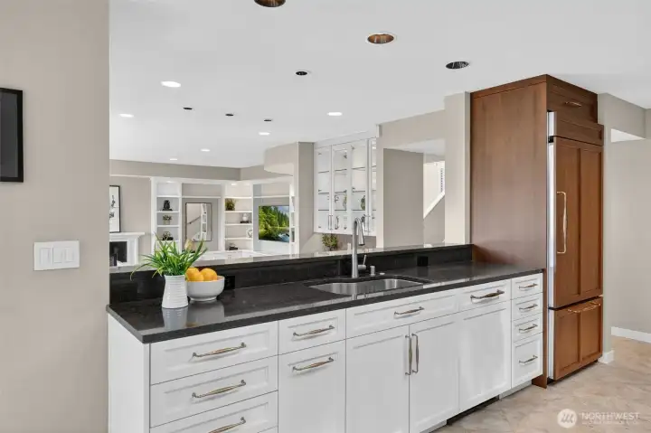 Dark granite countertops, brushed gold   hardware, and an undermount sink anchor   this working peninsula, while the   pass-through opens directly to the living   room and fireplace beyond — keeping the   cook connected to every conversation. The   scale of counter space and cabinetry here   reflects a kitchen designed for someone who   actually uses one.
