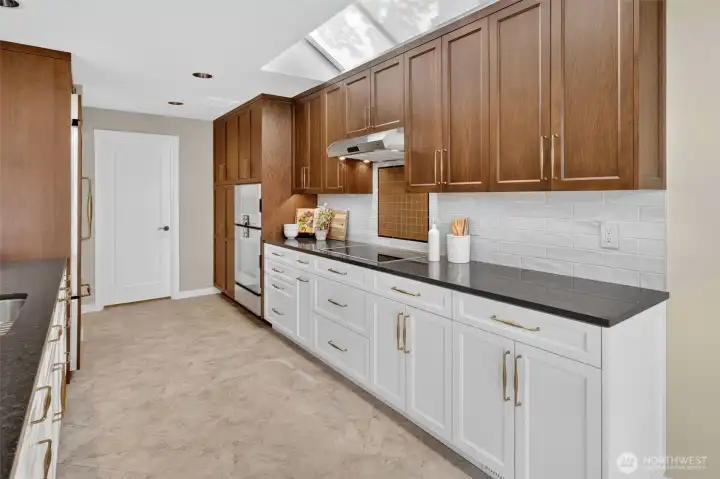 The full galley stretch reveals just how much   storage and prep space this kitchen truly   delivers — dual granite runs, gold hardware,   and a Gaggenau induction cooktop on one   side, undermount sink and panel-ready   refrigerator on the other. The door at the end   leads directly to the mudroom and garage,   keeping arrivals and departures seamlessly   tucked away from the main living areas