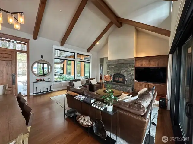 This inviting great room features vaulted ceilings with exposed beams, a floor-to-ceiling stone fireplace, and expansive windows for abundant natural light and seamless indoor-outdoor living.