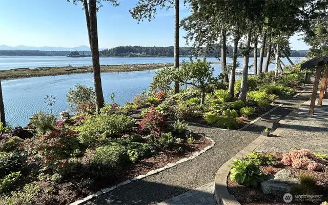 Winding paths draw you through a diverse array of flora, inviting leisurely strolls amidst flowering shrubs and verdant foliage, all backdropped by the tranquil waters and distant mountains.