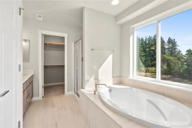 Soaking tub with tile surround in the primary bathroom, featuring a large window, a walk-in shower and direct access to the primary walk-in closet.