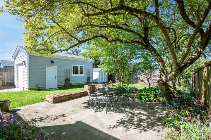 Looking toward the garage, this patio has a beautiful Magnolia Tree that acts as a canopy for shade from the SW Sun.   lawn with patio & garden spaces make this outdoor oasis easy to maintain & relax in.   Mature landscaping surrounds the home with established trees, shrubs, and seasonal blooms. Relax on the back patio under a green canopy all summer next to the outdoor stone fire pit. A dry, semi-finished basement offers flexible rec room / home gym space with fresh flooring.    Fully fenced property with 6' privacy fencing in back and black chain-link fencing with a remote-controlled rolling gate at the front. Deep driveway offers multiple off-street parking spots and leads to newer 14’x24’ detached garage with an extra tall ceiling offering generous storage options and accommodation for tall vehicles.    Roof and side sewer u