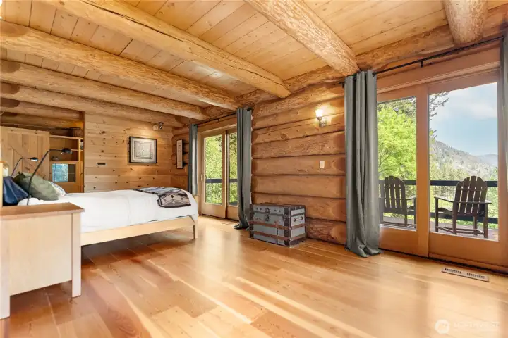 Main-floor primary suite with massive timber construction, custom window seat, walk-in closet, and sliding doors opening to a large private deck overlooking the surrounding mountain landscape. Warm natural light and handcrafted lodge details create a peaceful retreat experience.