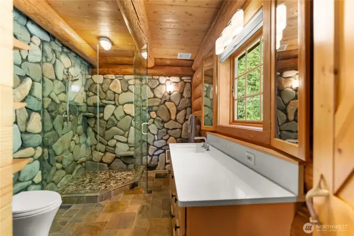 Light-filled primary bath featuring a custom river rock and glass shower, warm wood finishes, and artisan lodge-style details designed to reflect the natural beauty of the surrounding Methow landscape.