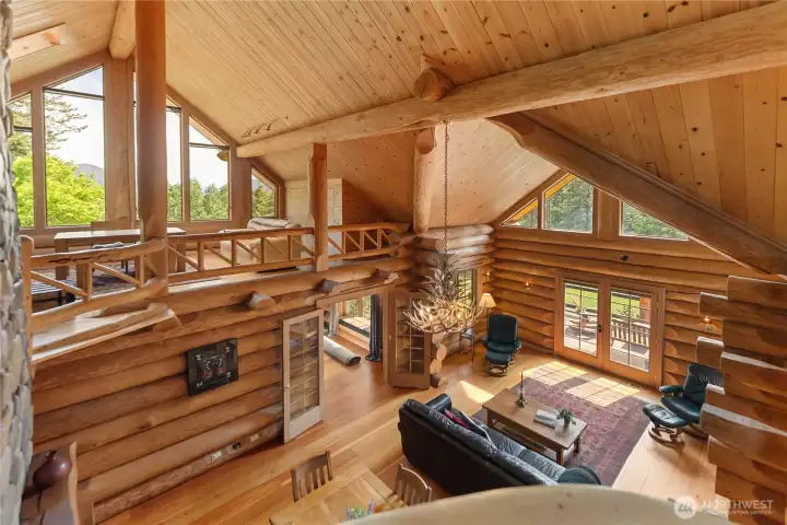 Vaulted great room filled with natural light, soaring wood ceilings, dramatic windows, and never-ending wood details that showcase exceptional craftsmanship throughout. Open loft above overlooks the living area, anchored by a striking river rock fireplace and warm custom finishes that frame the surrounding forested landscape.
