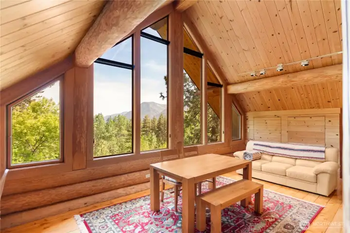 Dramatic wall of windows fills the loft with natural light and frames sweeping mountain and forest views, while soaring wood ceilings, exposed log beams, and warm natural finishes create a true mountain retreat atmosphere above the great room below.