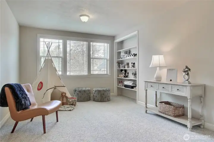 The upper wing bonus room is a bright and welcoming space, perfect for relaxing or entertaining. Built-in shelving keeps your favorite games, books, and cozy blankets neatly organized, making it easy to unwind or gear up for fun. Whether you're enjoying a movie night or diving into game time, the conveyed TV is ready for it all. Large windows fill the room with natural light by day, while the warm ambiance makes it just as inviting after sunset. It’s the perfect upstairs retreat for both quiet moments and lively gatherings.