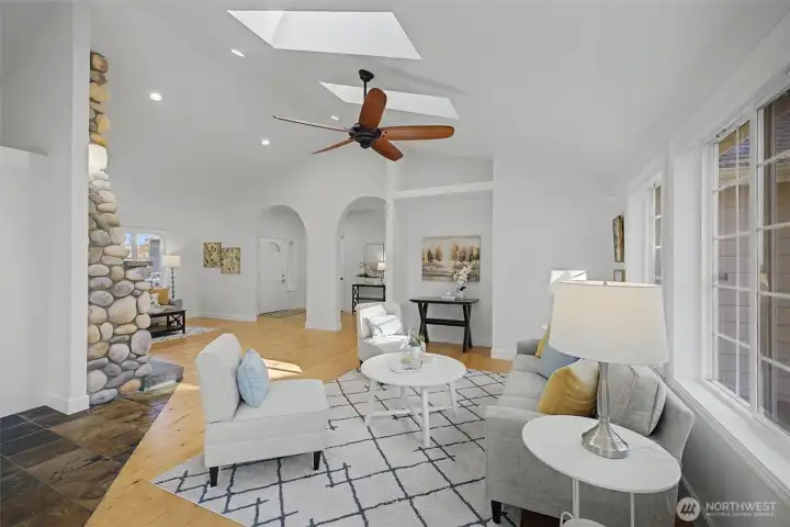 The vaulted great room is filled with natural light from dual skylights, recessed lighting and tall windows that create a bright, airy atmosphere. Warm wood floors, a modern ceiling fan and soft neutral finishes enhance the inviting seating area, while elegant arched openings frame views toward the foyer and adjoining living spaces. The dramatic floor-to-ceiling river rock fireplace adds character and warmth, anchoring this stunning central gathering space.