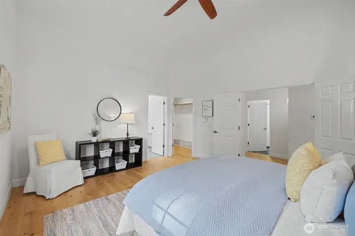 Spacious primary bedroom featuring vaulted ceilings, warm wood flooring and a soft neutral color palette that enhances the room’s light, open feel. Generous wall space easily accommodates a king-size bed and additional furnishings, while the open layout creates a sense of calm and flexibility. The walk-in closet and adjacent hallway access are thoughtfully positioned just off the bedroom for everyday convenience