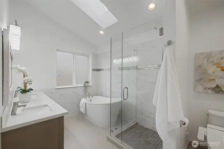 Bright and beautifully updated primary bathroom featuring a freestanding soaking tub, a frameless glass walk-in shower and elegant marble-style wall tile with a decorative accent band. A skylight and recessed lighting enhance the airy feel, while the large window brings in soft natural light. The double-sink vanity with quartz countertops and modern fixtures adds both style and functionality, creating a well-balanced and thoughtfully designed space.