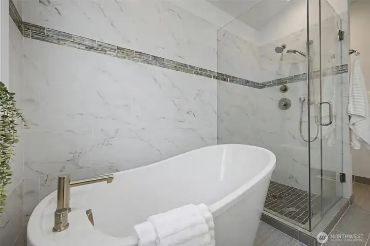 Close-up view of the primary bathroom highlighting the sculptural freestanding soaking tub set against marble-style tile walls with a sleek accent inlay. The adjacent frameless glass shower features modern fixtures and a tiled shower floor for a clean, cohesive look. Soft neutral finishes, crisp white surfaces and contemporary hardware combine to create a polished, tranquil atmosphere with a refined architectural feel.
