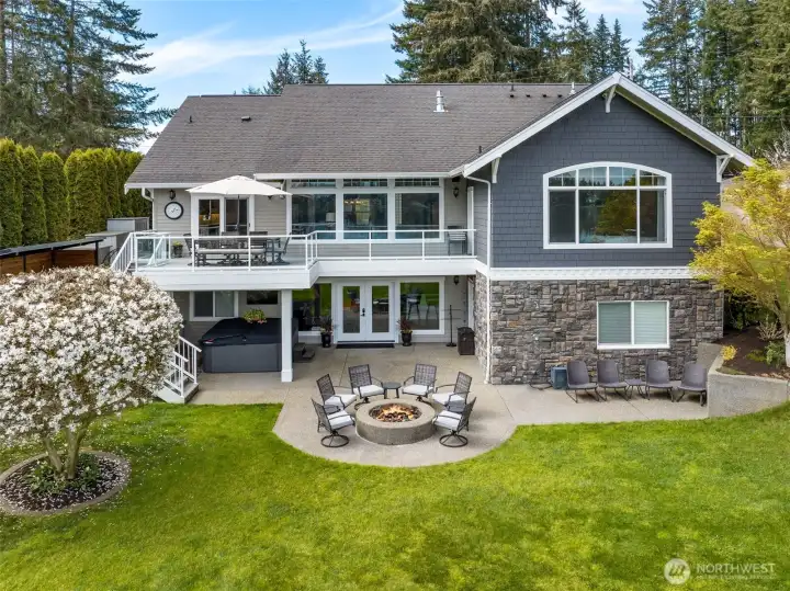 The extended patio gives you real space to spread out, not just a token slab, and it flows effortlessly into the lawn and up to the deck above. That flowering tree adds a pop of softness and color that makes the whole setting feel a little more special without trying too hard. The stone detailing along the lower exterior grounds the home beautifully, while that arched window in the primary suite quietly steals the show. Everything feels connected, usable, and thoughtfully done.