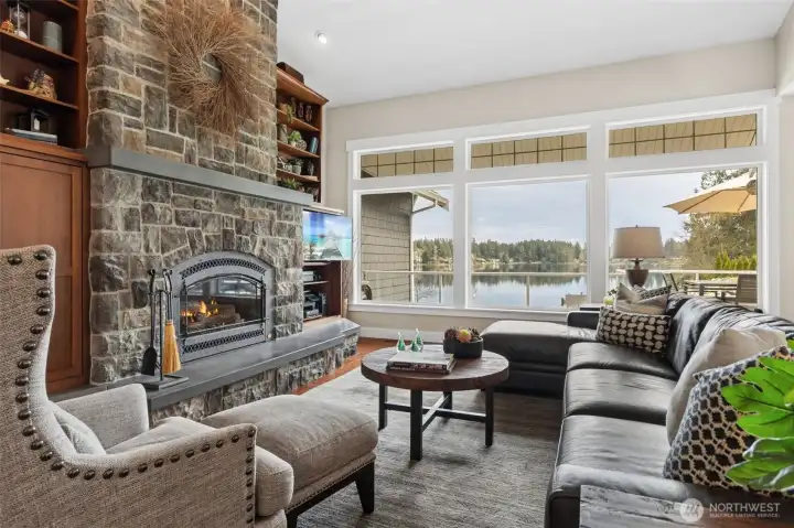 This is the room that earns the “wow.” Three oversized windows line up across the back, framing the glass-railed deck and Long Lake like it’s on display, and those upper accent windows layer in even more light without interrupting the view. Front and center, the floor-to-ceiling stone fireplace brings texture and presence, blending soft grays with warmer sandstone tones that play perfectly against the creamy walls. Built-in shelving on both sides keeps things balanced and functional without stealing attention.