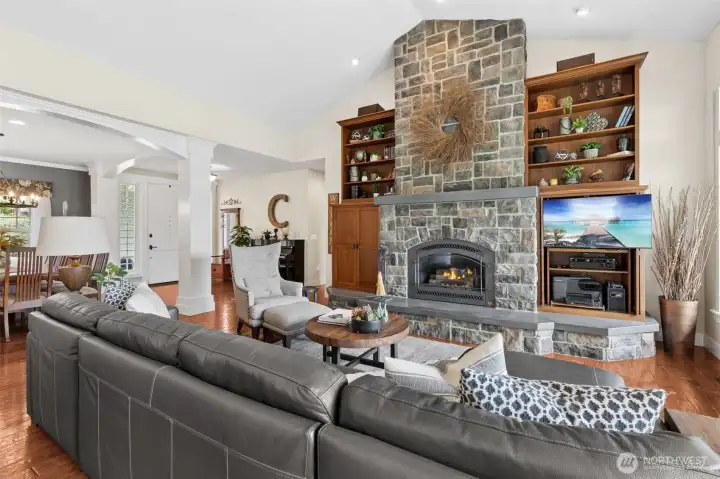 From this corner, the room really shows off its scale without losing that cozy factor. The fireplace stretches fully into view, complete with a long hearth that runs across the built-ins, giving it a grounded, almost lounge-like feel. It’s the kind of space where you can actually settle in, not just admire. Up top, you’ll notice the integrated speakers, part of a whole-home sound system that carries both inside and out, because ambiance matters. The architectural details start to layer in here too, pillars and that arched opening subtly guide you toward the entry and formal dining without closing anything off. At over 4,000 square feet, this home knows how to give you space while still feeling inviting.