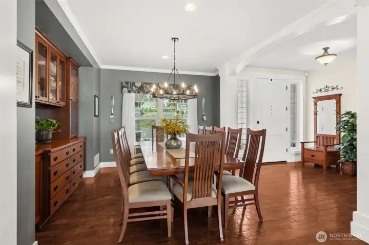 Right out of the gate, this home makes it clear it’s not playing small. The entry opens with wide plank hickory floors that carry warmth and presence, flanked by accent windows that pull in natural light without sacrificing privacy. There’s room here to land, a bench, a statement console, something that says “you’ve arrived.” And then the dining room steps in and takes over. A chandelier that actually earns its spot, stately gray walls with crisp crown molding, and enough space to host a full table for twelve without anyone bumping elbows. The built-in buffet with glass-front cabinetry is both practical and a little showy, in a good way.