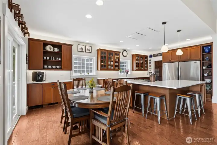 Natural light pours in from every angle, sliding glass doors, multiple windows, even a peek of those character-filled panes over the sink and desk area. There’s a full casual dining zone that comfortably handles a table for six, plus a massive island that pulls double duty for prep and seating. Quartz counters keep it clean and bright, while the warm-toned cabinetry and soft cream walls make the whole space feel grounded and inviting. Storage is dialed in with pullouts where you actually need them, a mix of closed cabinetry, glass-front uppers, and open shelving that keeps things from feeling heavy. And then there’s the flex you didn’t know you needed, two full refrigerator/freezer combos and a floor-to-ceiling display wall that’s ready for everything from art pieces to everyday essentials.