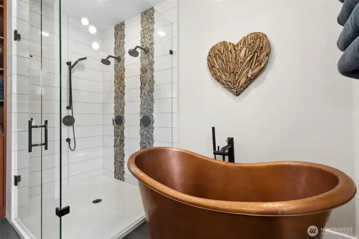 The hammered copper vessel tub steals the spotlight in the best way, rich, sculptural, and not something you see every day. Matte black fixtures ground the space and keep it feeling current, while the glass-enclosed shower brings the spa energy with three showerheads and a striking floor-to-ceiling river rock accent. It’s detailed without feeling busy. The low-threshold entry makes it easy to step in and out, adding a practical layer to the design, and the slate-toned tile floors tie everything together with a clean, natural finish.