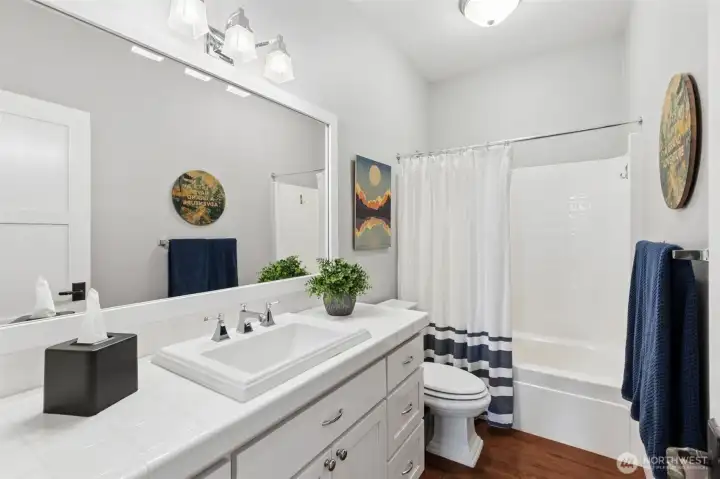 You’ve got 9-foot ceilings carrying that same open feel from the rest of the main level, paired with a full tub and shower setup that’s ready for everyday use. The vanity stays crisp and clean with white shaker cabinetry, tile countertops, and chrome fixtures that keep everything feeling fresh. And then there’s that integrated porcelain surround at the sink and toilet, a molded base, which adds a subtle, almost custom detail you don’t see very often. Hickory floors continue through here, tying it back to the rest of the home and keeping the look cohesive.