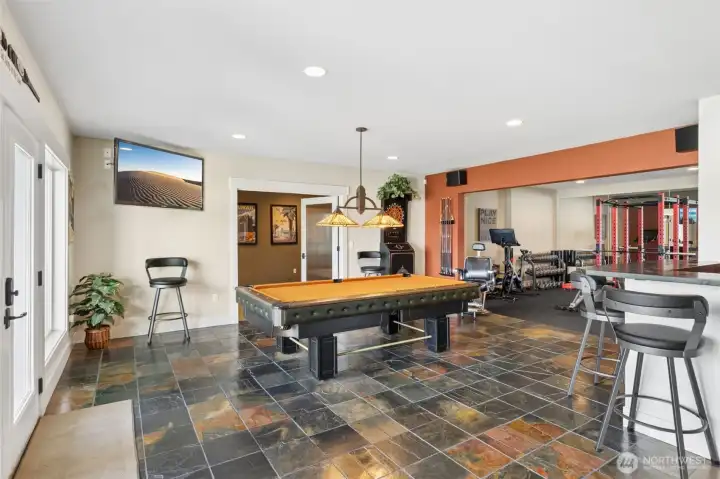 Here’s where you really understand the layout. Every zone has its place, but nothing feels boxed in. The bonus room is just beyond those glass-paneled doors, the gym reflects light and energy back into the space, and the bar anchors the social side of things. It all connects in a way that feels easy and natural. The slate floors tie everything together with a mix of deep and warm tones that give the room character without overwhelming it.