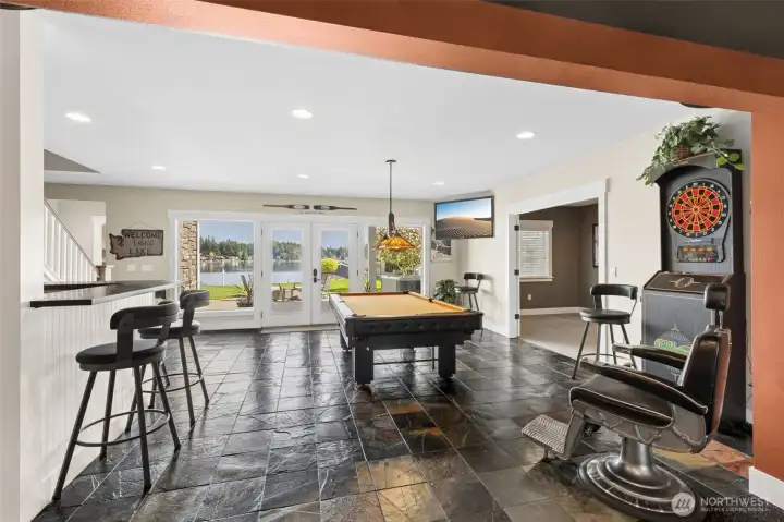 Stand in the gym and suddenly the whole lower level unfolds. Straight ahead, French doors framed by two oversized windows pull your eye right out to the patio and lake beyond, so even down here, you’re still connected to the view. To the right, those reeded glass doors lead into the bonus room, giving you that extra flex space without closing things off. To the left, bar seating with beadboard detail quietly says “yes, this level is meant to be used.” And tying it all together, those warm, creamy tones keep the space from feeling like a basement at all.