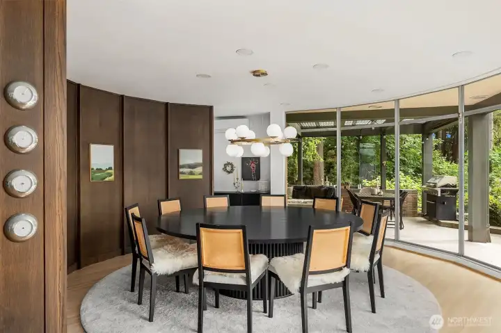 Circular dining room with solid wood paneling and floor to ceiling windows towards covered patio area. Circular dining room with solid wood paneling and floor to ceiling windows towards covered patio area.