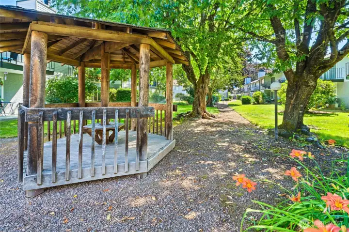 Rustic gazebo surrounded by shade trees and flowers provides lovely outdoor gathering space for community.
