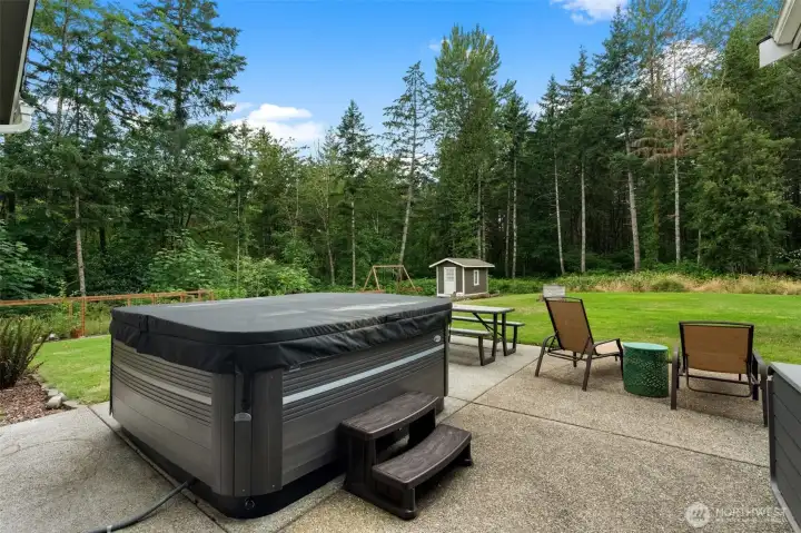 Covered patio features a hot tub, seating area, and views of the expansive lawn and surrounding trees.