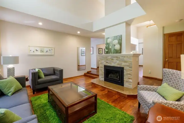 As you reach the end of the hallway, there's a statement fireplace. Step down into the living room and find high, vaulted ceilings, wall of windows, engineered hardwood floors giving a lush garden feeling to this room and orienting towards the gardens.