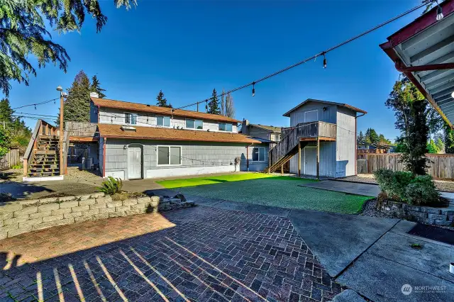 Fully fenced back yard patio and play area with artificial grass and an out building.