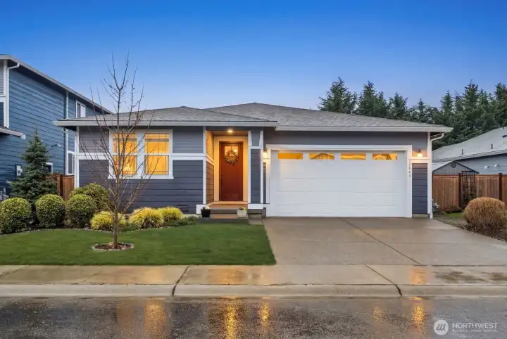Twilight exterior showcasing the Bonney Lake rambler with covered front entry, illuminated interior windows, attached two-car garage, and landscaped front yard along a residential street in the Falling Water neighborhood.