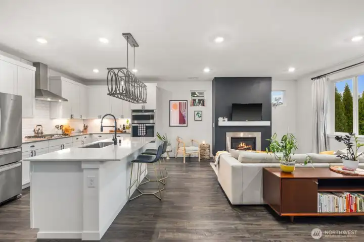 Open concept living space featuring a large white kitchen island with seating, white cabinetry, stainless steel appliances, recessed lighting, and a living area with fireplace and large windows providing natural light.