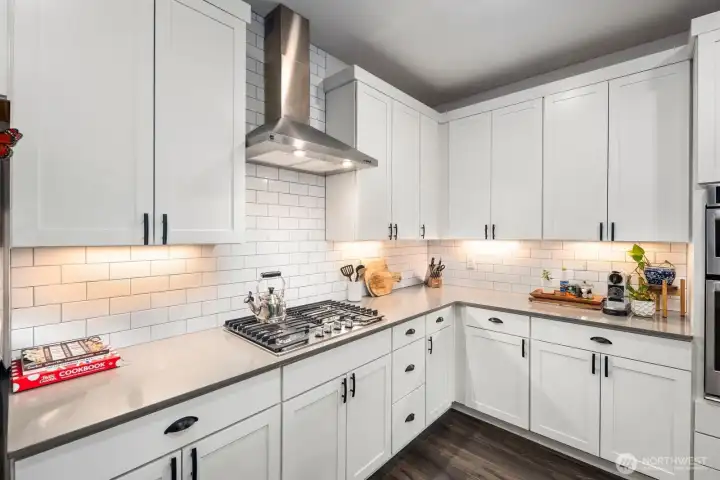 Kitchen corner featuring white cabinetry, subway tile backsplash, stainless steel vent hood, gas cooktop, and under-cabinet lighting illuminating the workspace.