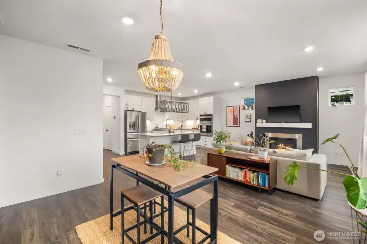 Wide interior perspective capturing the living room, kitchen, and adjacent dining area, emphasizing the open layout and natural light from large windows.