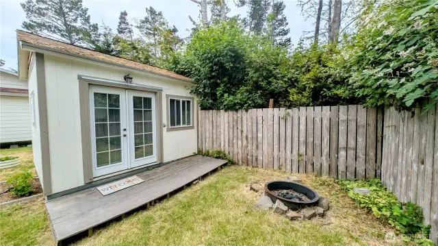 This storybook backyard retreat blends rustic charm with relaxed outdoor living. A welcoming cottage with French doors and a wooden deck invites creativity, rest, or overnight guests, while the nearby firepit sets the scene for cozy gatherings under the stars. Framed by trees, fencing, and a soft lawn, it’s a private haven perfect for s’mores, storytelling, and slow mornings with coffee in hand