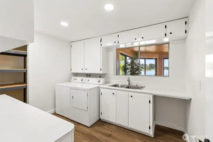 Leave it to a waterfront home to give even the laundry room a view. This spacious utility room looks through an interior window into the lake-facing lower sunroom, creating a bright and surprisingly cheerful workspace that feels connected to the outdoors. A utility sink sits perfectly positioned beneath the window, while abundant cabinetry, long counters and dedicated storage areas provide room for everything from laundry supplies to craft projects and holiday decorations. Waterproof luxury vinyl plank flooring stands up beautifully to everyday use, and recessed lighting keeps the entire space feeling fresh and modern. Leave it to a waterfront home to give even the laundry room a view. This spacious utility room looks through an interior window into the lake-facing lower sunroom, creating a bright and surprisingly cheerful workspace that feels connected to the outdoors. A utility sink sits perfectly positioned beneath the window, while abundant cabinetry, long counters and dedicated storage areas provide room for everything from laundry supplies to craft projects and holiday decorations. Waterproof luxury vinyl plank flooring stands up beautifully to everyday use, and recessed lighting keeps the entire space feeling fresh and modern.