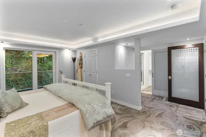 The serene primary suite is thoughtfully positioned at the rear of the residence, overlooking a lush, verdant hillside. It features a private balcony with sleek glass panels, offering a peaceful outdoor retreat. Inside, a tray ceiling with integrated lighting adds depth and elegance, enhancing the room’s calm and refined atmosphere.