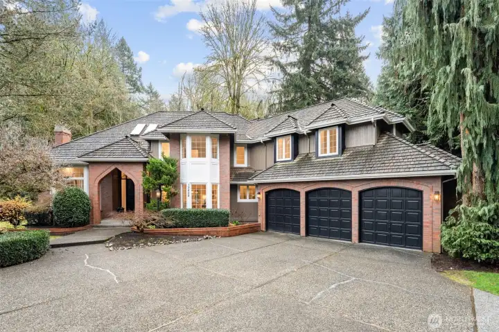 A stately, timeless exterior with classic brick accents, mature evergreens, and a wide driveway leading to a spacious three-car garage—an elegant first impression nestled in a serene, wooded setting.