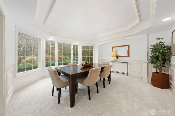 Elegant dining room featuring a tray ceiling, classic wainscoting, and expansive windows that frame lush greenery—an inviting space designed for memorable gatherings and refined meals.