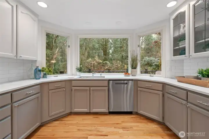 Bright kitchen workspace with quartz counters, stainless steel dishwasher, and oversized windows overlooking lush greenery—bringing natural light and a serene backdrop into the heart of the home.