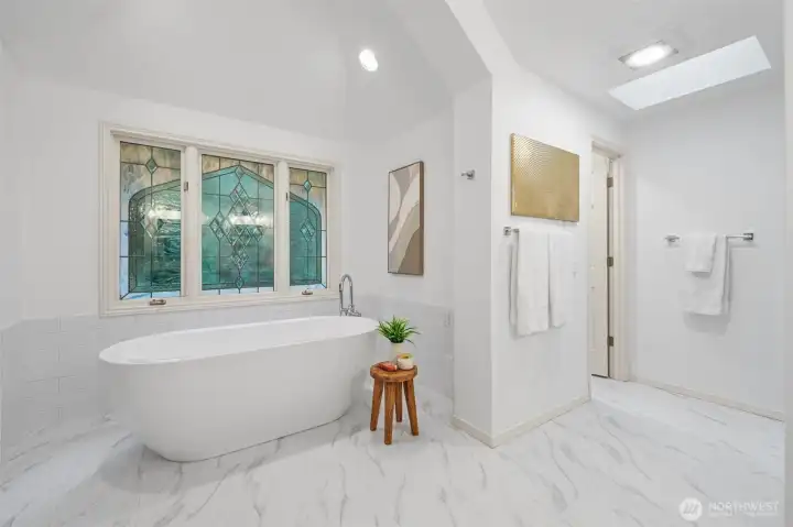 The luxurious soaking area showcases a freestanding tub set beneath a stunning decorative window, complemented by marble-style flooring and a nearby walk-in shower—perfect for relaxation and everyday comfort.