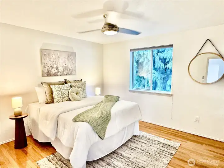 Bright and airy bedroom with beautiful hardwood floors, a ceiling fan, and large windows overlooking mature landscaping.