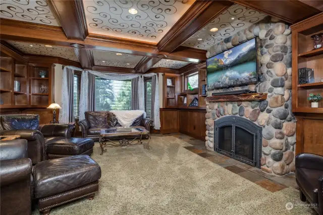 The library features elaborate ceiling inlays, exposed beams, a river rock gas fireplace, and extensive built-in shelving.  The flooring is carpet, and there is a bay window