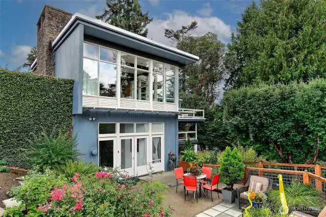 Viewed from the west garden, the home’s vertical drama comes into focus—floor-to-ceiling windows soaring across two levels, framed in deep eaves and cobalt stucco. French doors open onto a private patio ringed by roses and hedges, creating a sunlit courtyard for play, planting, and weekend gatherings with room to spare.