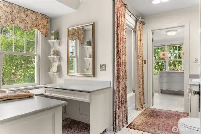 Light-filled upstairs bathroom with charming vintage details, dual vanities, and classic built-in shelving. Timeless design with floral accents, curated shelves, and a private dressing area overlooking the trees.